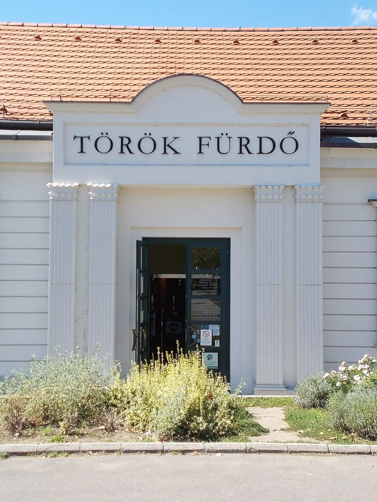 Façade blanche à pilastres sous un toit de tuiles orange, surmontée de l’inscription « TÖRÖK FÜRDŐ » ; la porte vitrée est ouverte, des massifs fleuris bordent le trottoir.